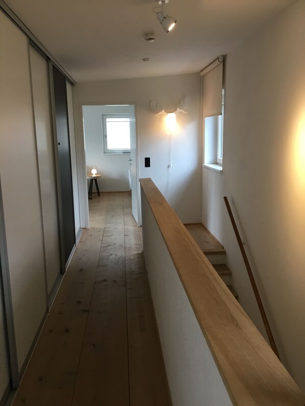 A narrow hallway with wooden flooring, a wooden handrail, and a window at the end.