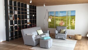 Ein Wohnzimmer mit grauem Sofa, grauem Sessel, Bücherregal und einem Gemälde von Mädchen in Kleidern.