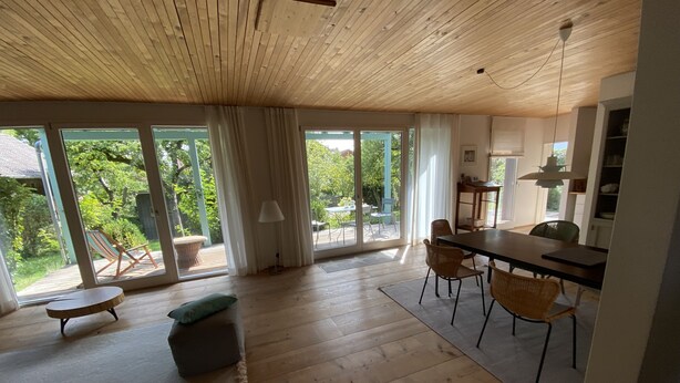 Ein geräumiges Zimmer mit Holzbalkendecke, großen Schiebeg lass-Türen, die auf eine Terrasse führen, einer Essecke mit Tisch und Stühlen und einer gemütlichen Sitzecke mit Sofa und Kissen.