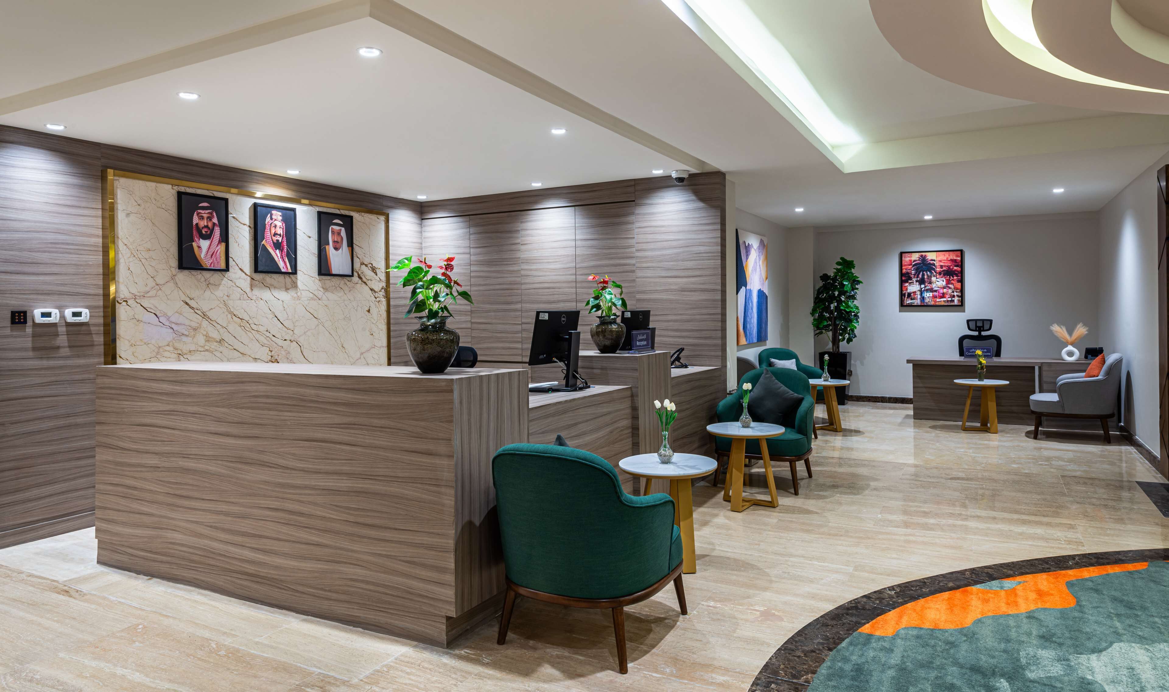 Un área de recepción moderna con un mostrador de mármol, varias opciones de asiento que incluyen un sillón verde y una pequeña mesa redonda, además de un mostrador con dos computadoras.