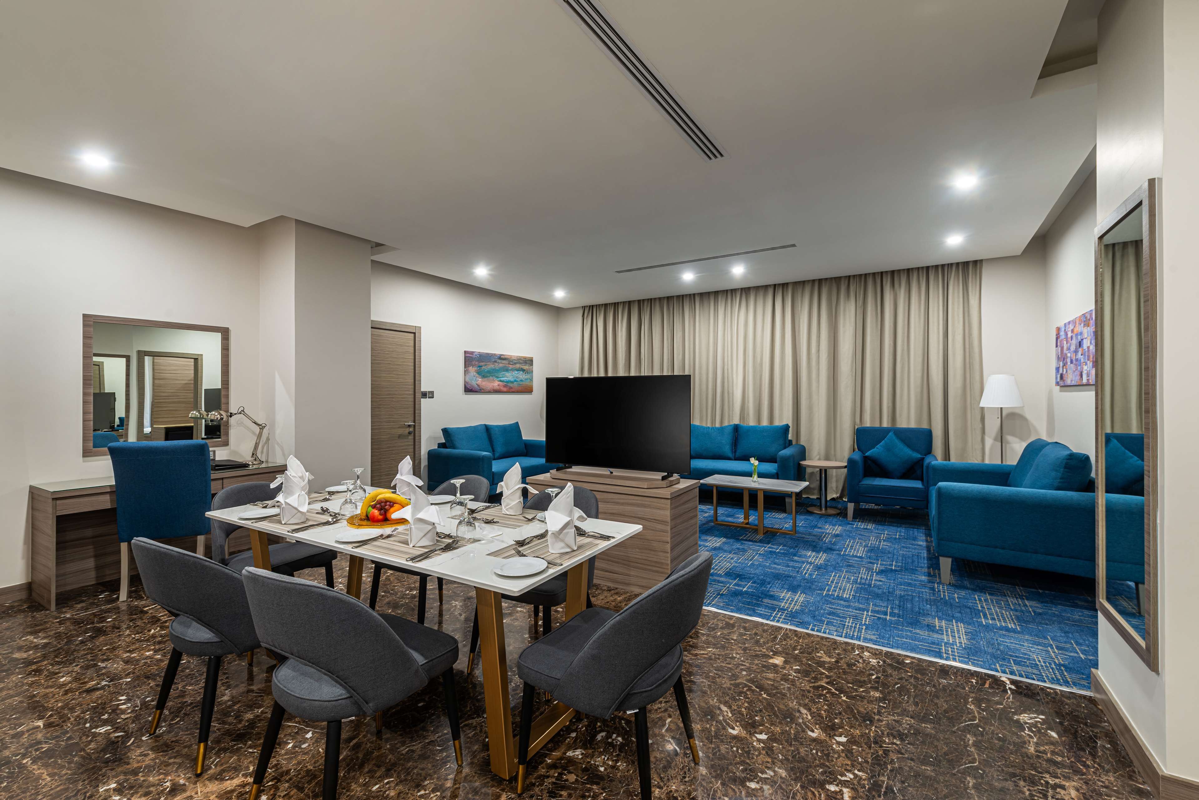 Een hotelkamer met een eetgedeelte, een woonkamer met blauwe banken, een flatscreen televisie en een groot raam met gordijnen.