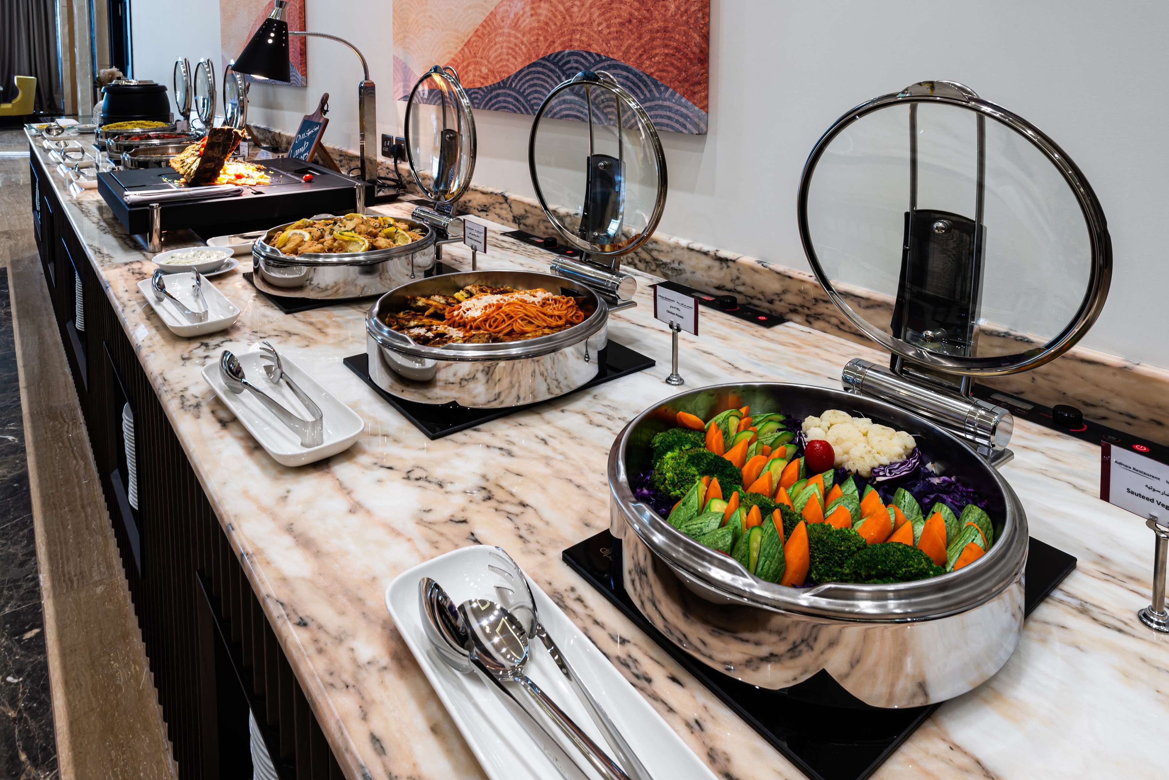 Buffet com diversos pratos, incluindo massas, legumes e carnes, dispostos sobre um balcão de mármore.