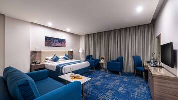 A hotel room with a bed, two blue sofas, a small table with a tray, a desk with a chair, a TV, and a large window with curtains.