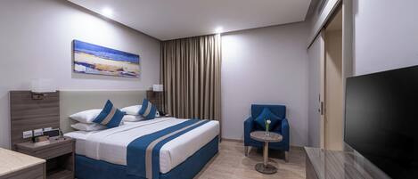 Una habitación de hotel moderna con una cama grande, un televisor de pantalla plana, una mesita redonda y un sillón azul.