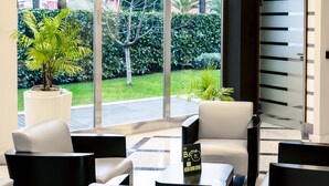 A modern indoor seating area with white leather chairs and black tables, overlooking a well-maintained garden with various plants and a tree.