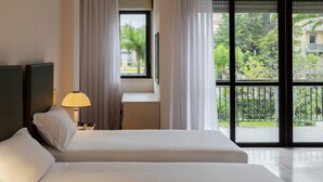 A hotel room with two beds, a nightstand with a lamp, a window with curtains, and a view of greenery outside.