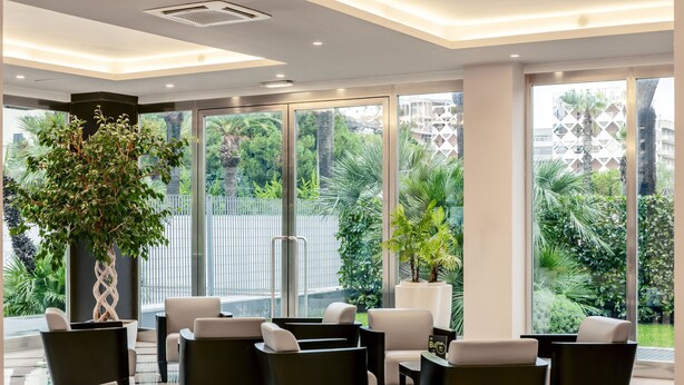 A modern lobby with a checkered floor, black and white armchairs, a large potted plant, and large windows offering a view of greenery and buildings.