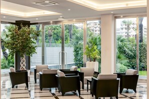 A modern lobby with a checkered floor, black and white armchairs, a large potted plant, and large windows offering a view of greenery and buildings. - B&B Hotel Bari Rondò (Bari)