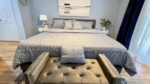 A bedroom with a bed, a bedside table, a lamp, a clock, and a window with curtains. - Tybee Island Silver Ave Retreat (Tybee Island)
