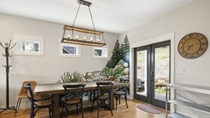 Un comedor con mesa y sillas de madera, una araña, un reloj y un mural con árboles y rocas en la pared.