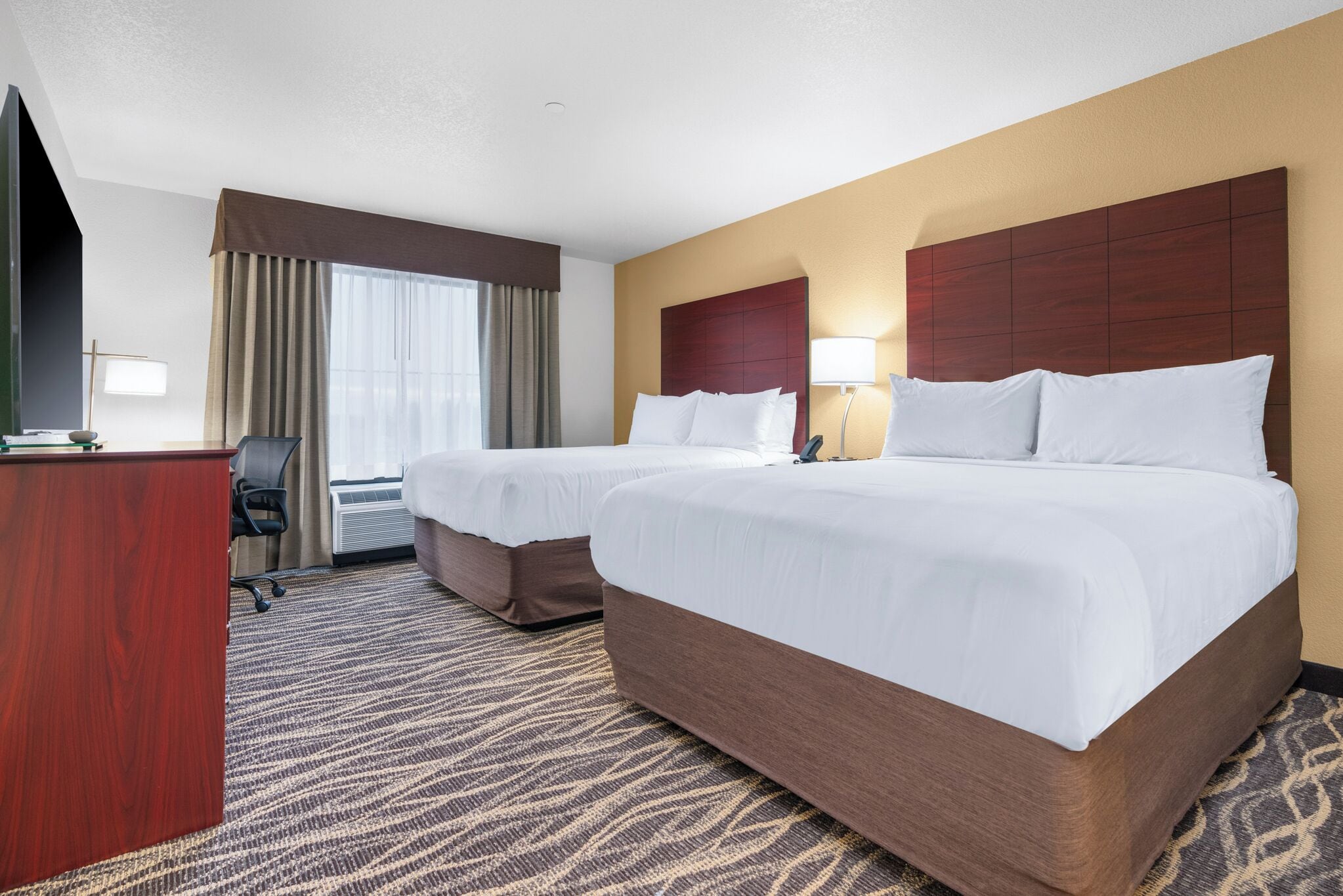 A hotel room with two beds, a desk, a chair, a lamp, and a window with curtains.