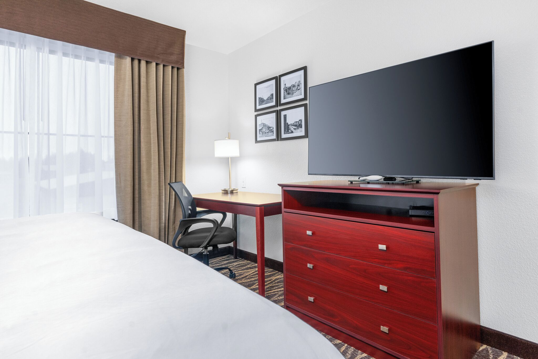 Une chambre d’hôtel comprenant un lit, un bureau, une chaise, une télévision, une commode et une fenêtre avec des rideaux.