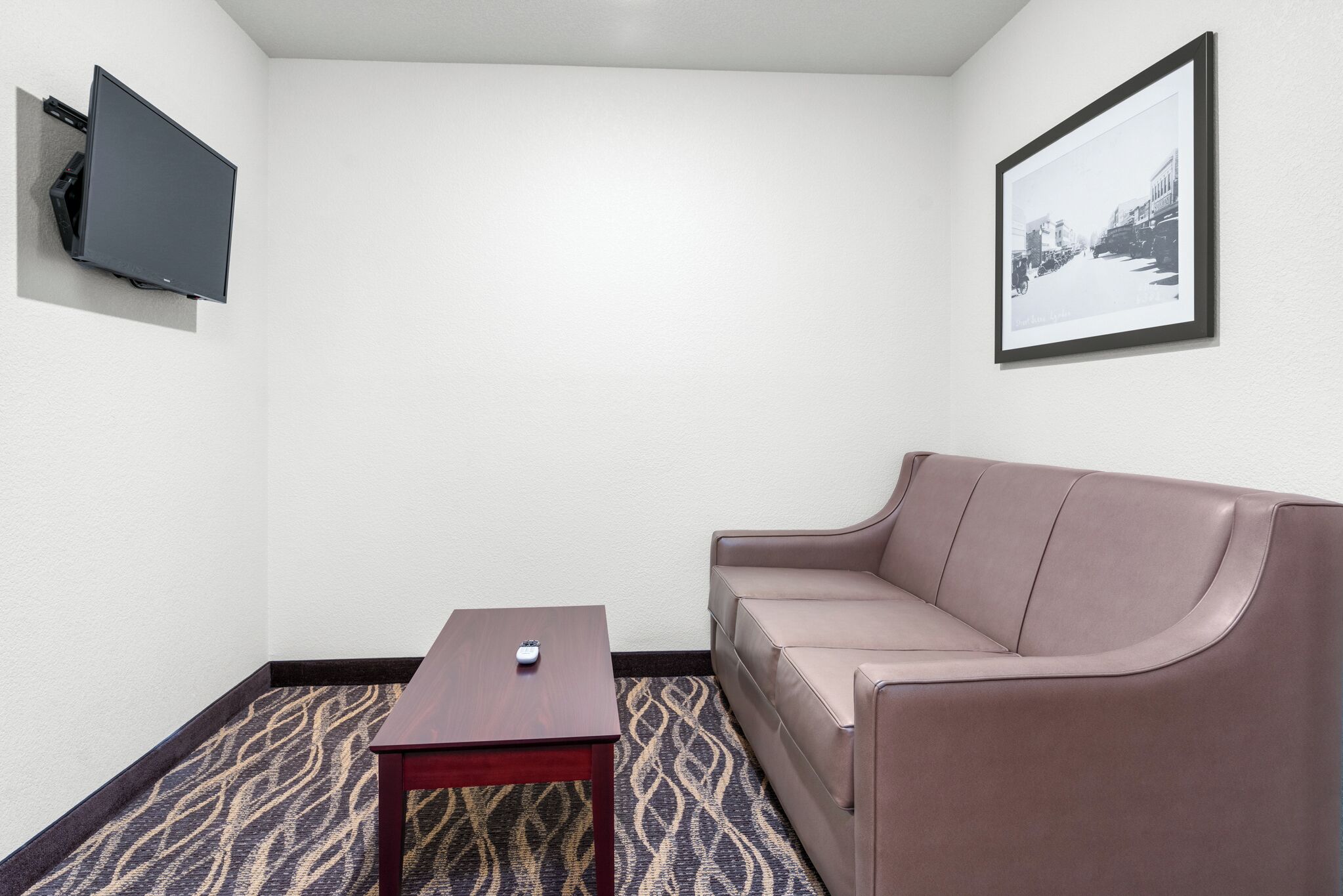 Una habitación con un televisor de pantalla plana montado en la pared, un sofá de cuero marrón, una pequeña mesa de madera y una alfombra estampada.