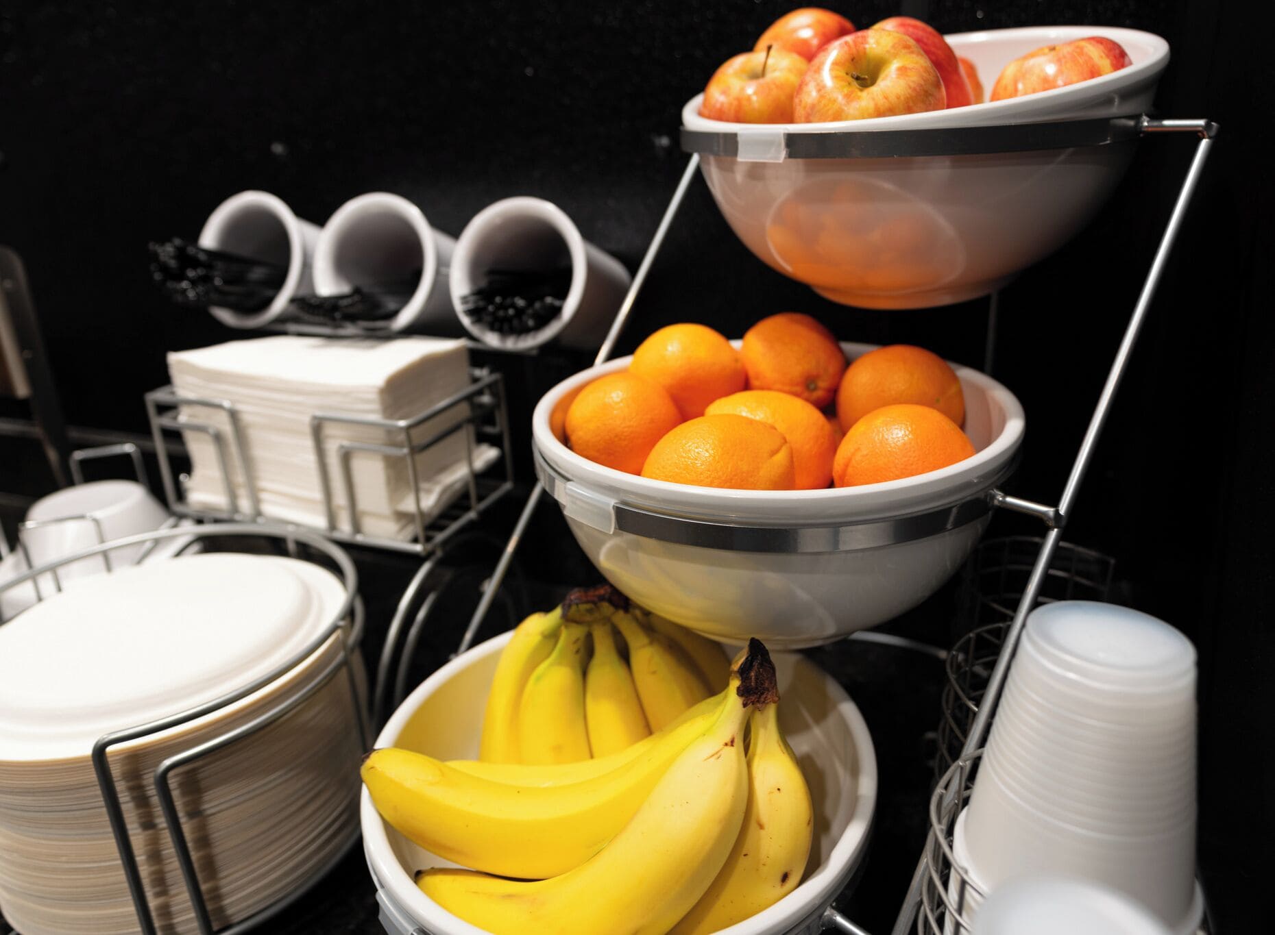 Ein Buffet mit Schalen voller Bananen, Orangen und Äpfeln sowie einem Stapel weißer Tassen.