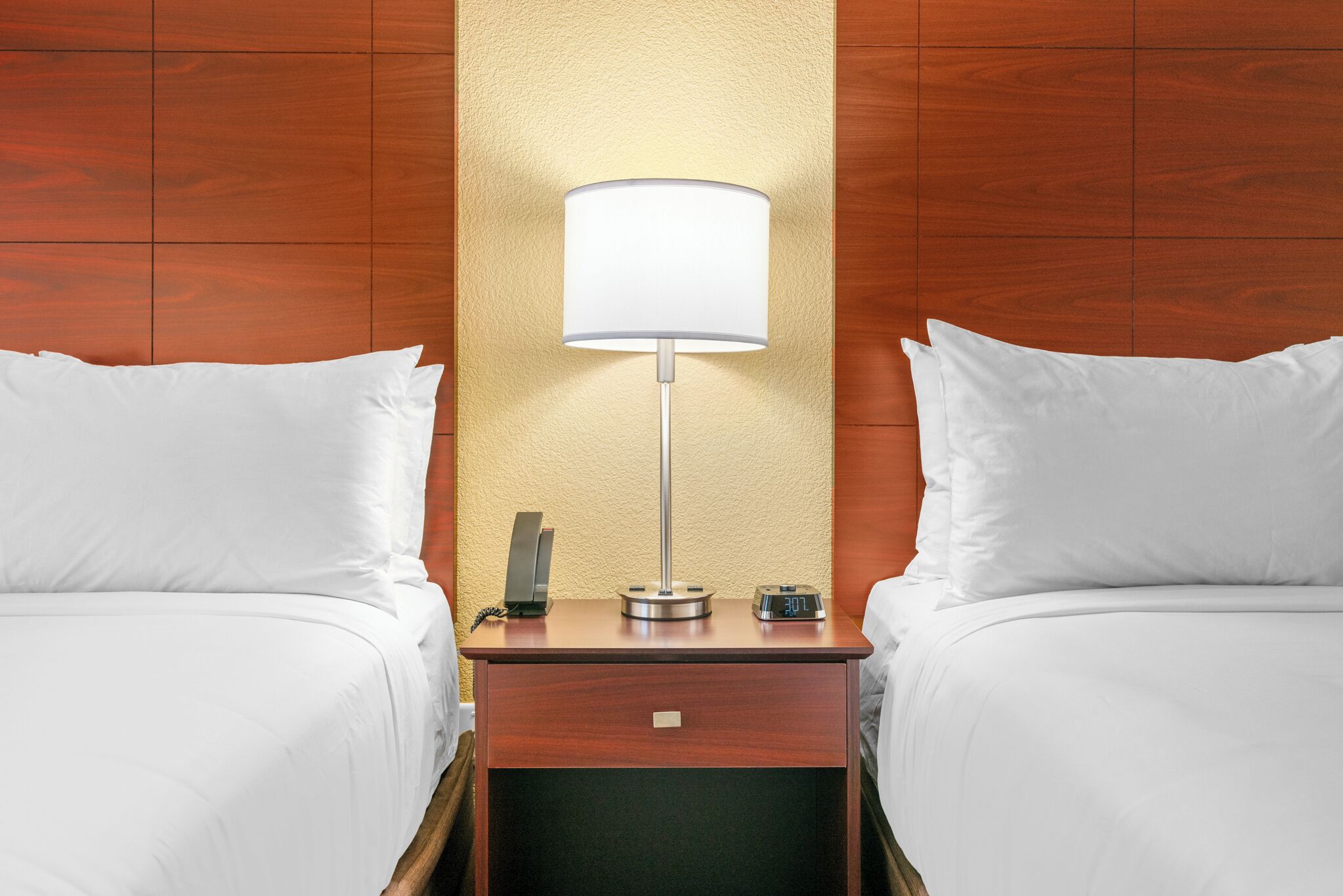 A hotel room with two beds, a nightstand with a lamp, a clock, and a phone.