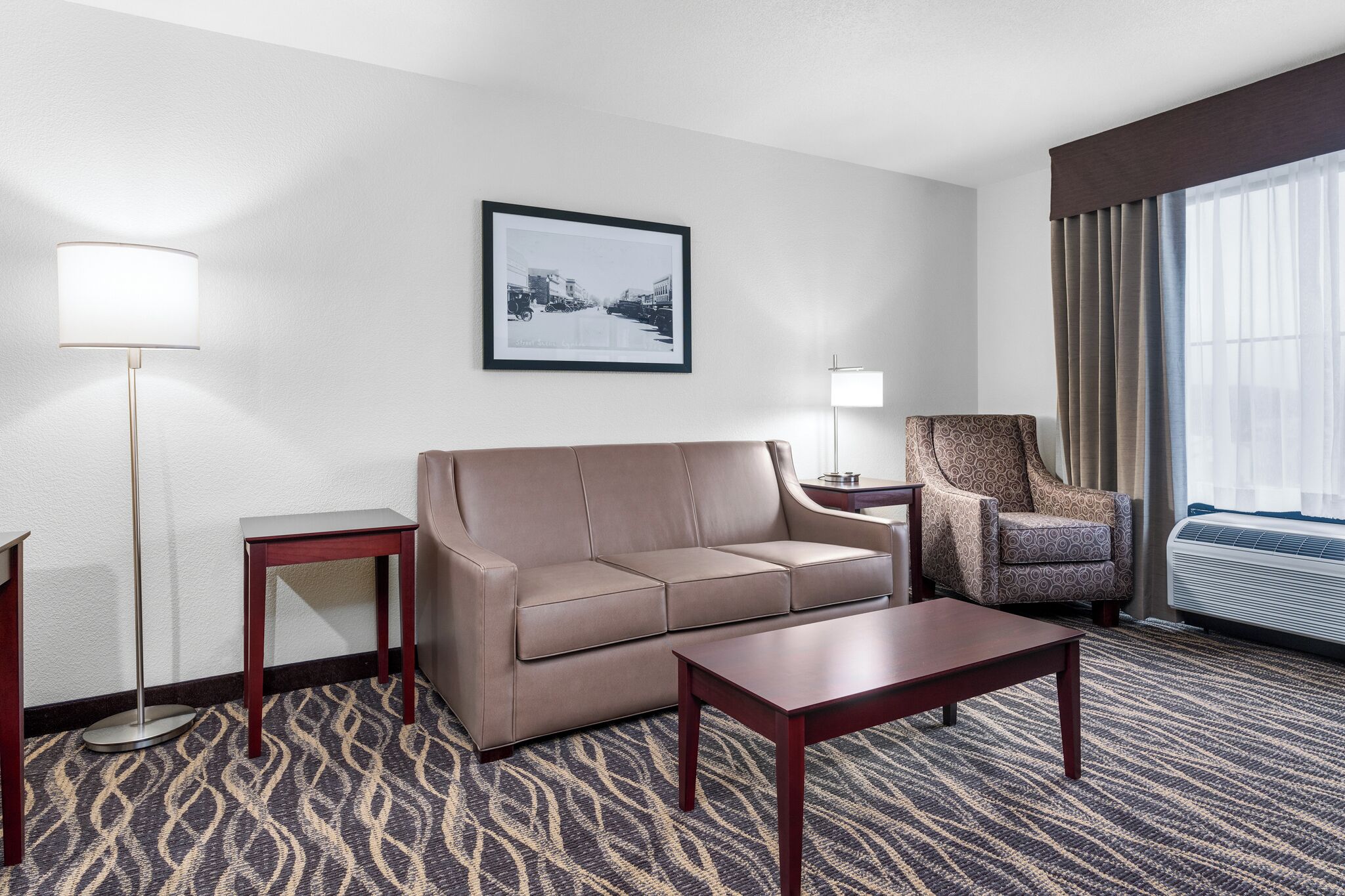 A hotel room with a sofa, armchair, two side tables, a lamp, and a framed picture on the wall.