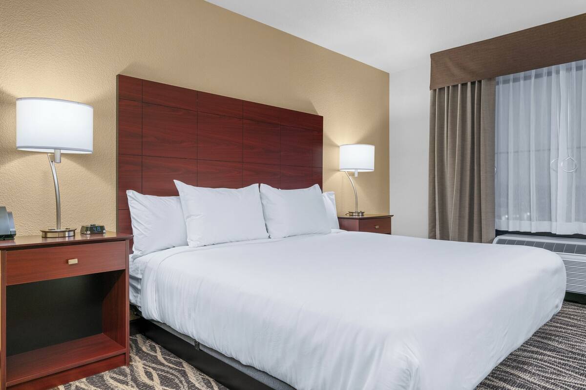 A hotel room with a large bed, two bedside tables, a nightstand, a wall-mounted lamp, and a window with curtains.
