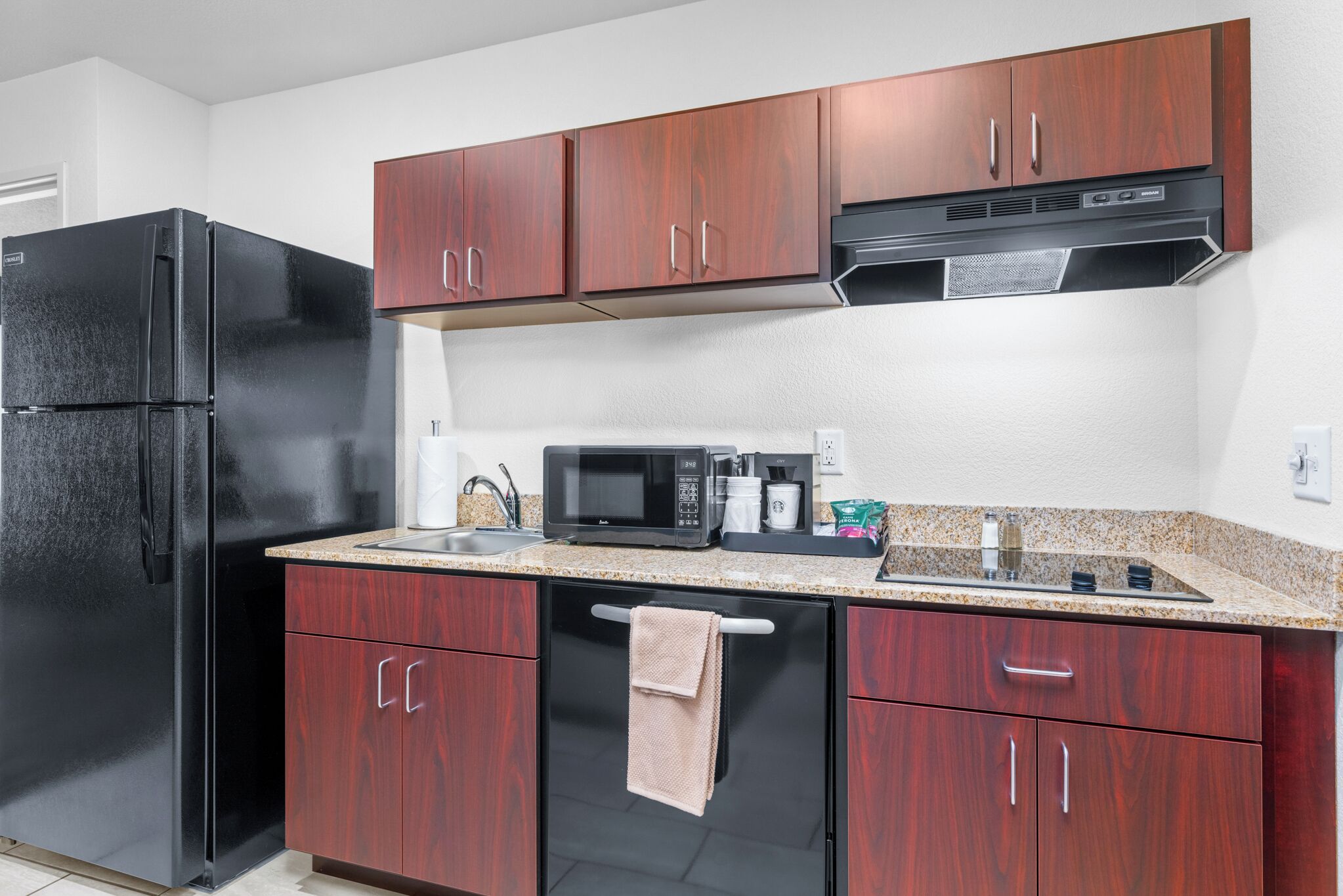 Una cocina compacta con un refrigerador negro, armarios de madera, un microondas y una cafetera.