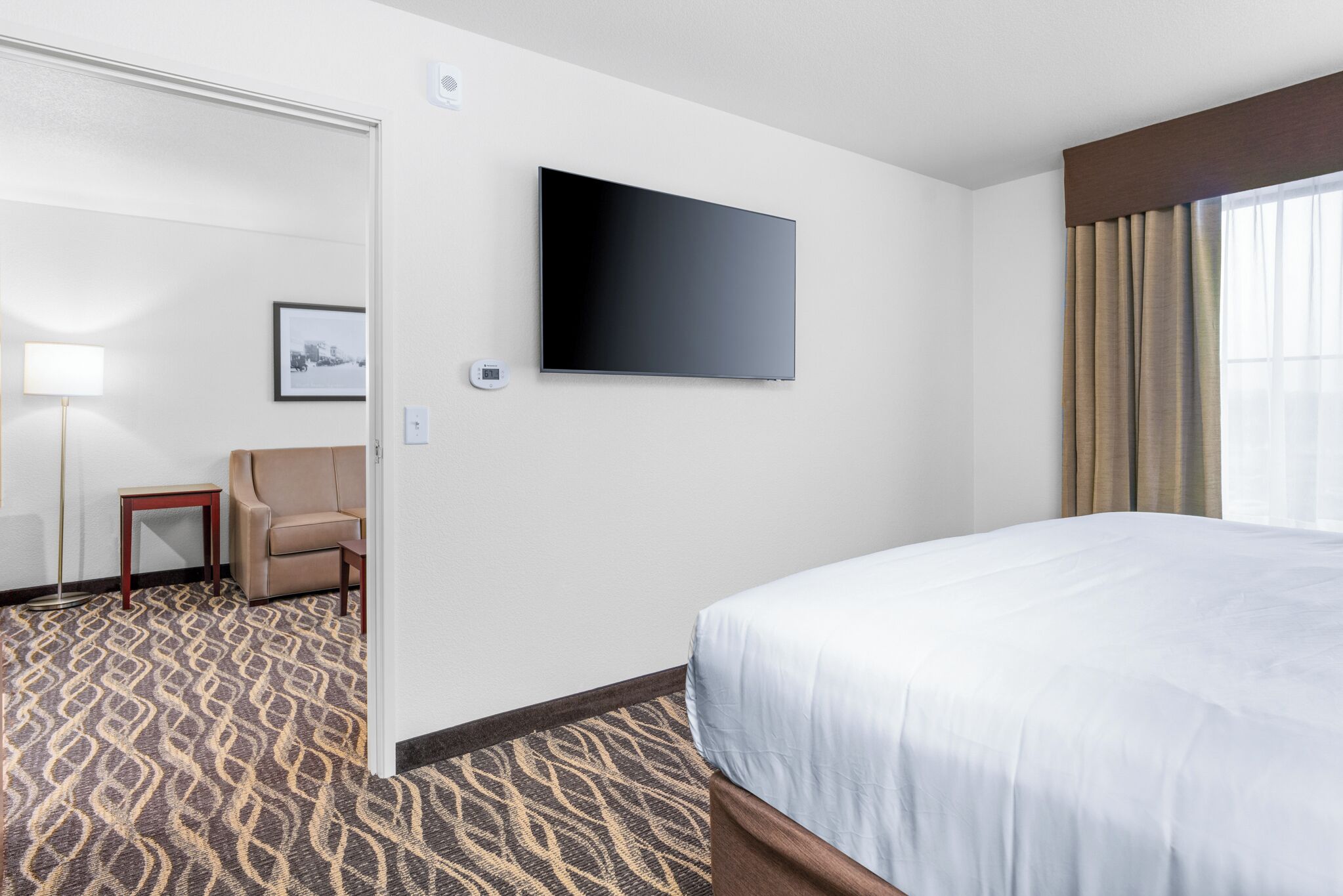 A hotel room with a large bed, a flat-screen TV mounted on the wall, a small table with a lamp, a chair, and a patterned carpet.