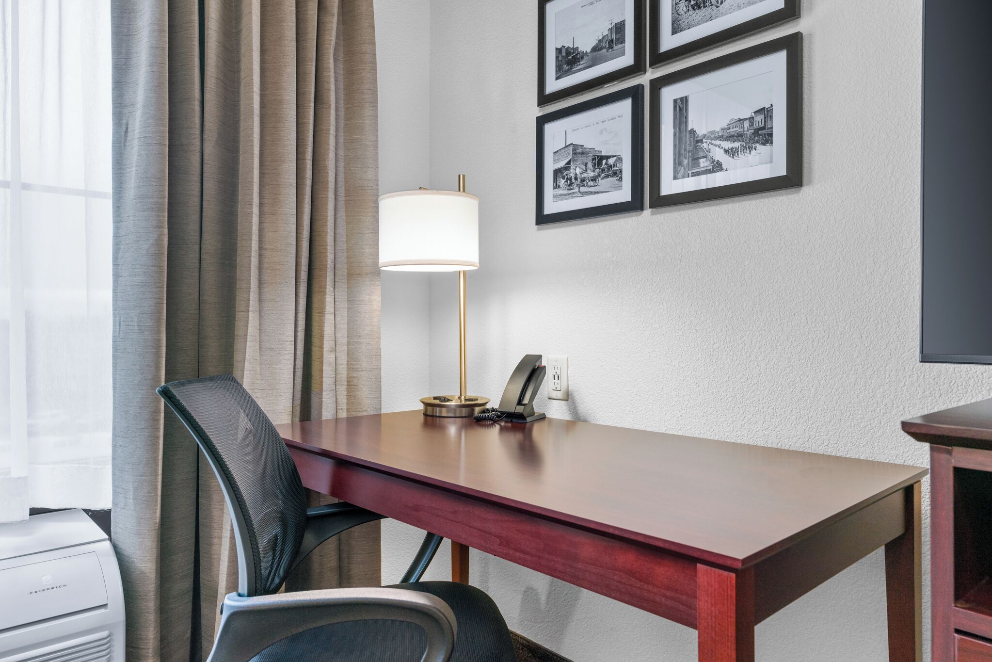 Habitación de hotel con un escritorio de madera, una silla, una lámpara y cuadros en la pared.