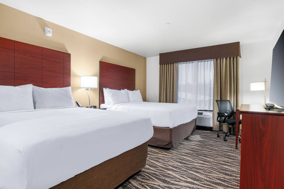 A hotel room with two beds, a desk, a chair, a lamp, and a window with curtains.