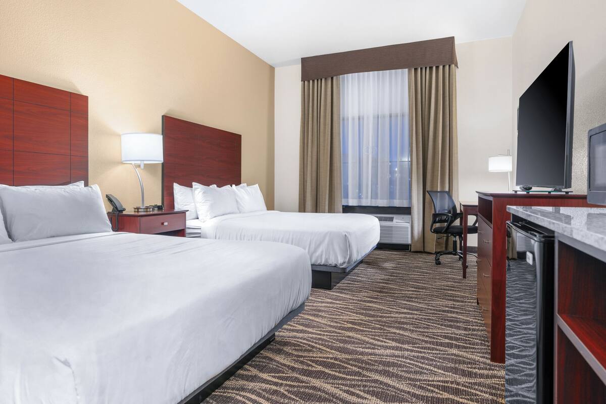 A hotel room with two beds, a desk with a TV, a desk lamp, a chair, and a window with curtains.