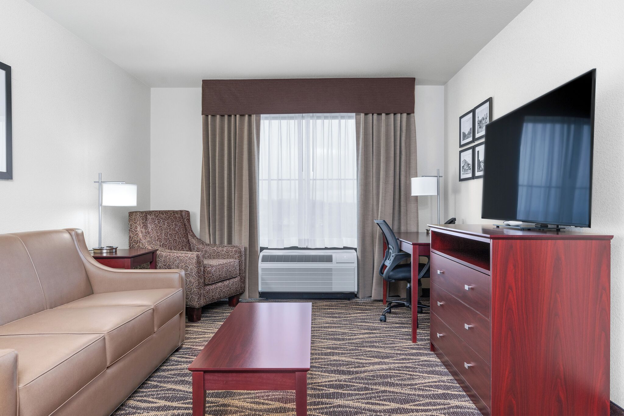A hotel room with a sofa, chair, desk, TV, and air conditioning unit.