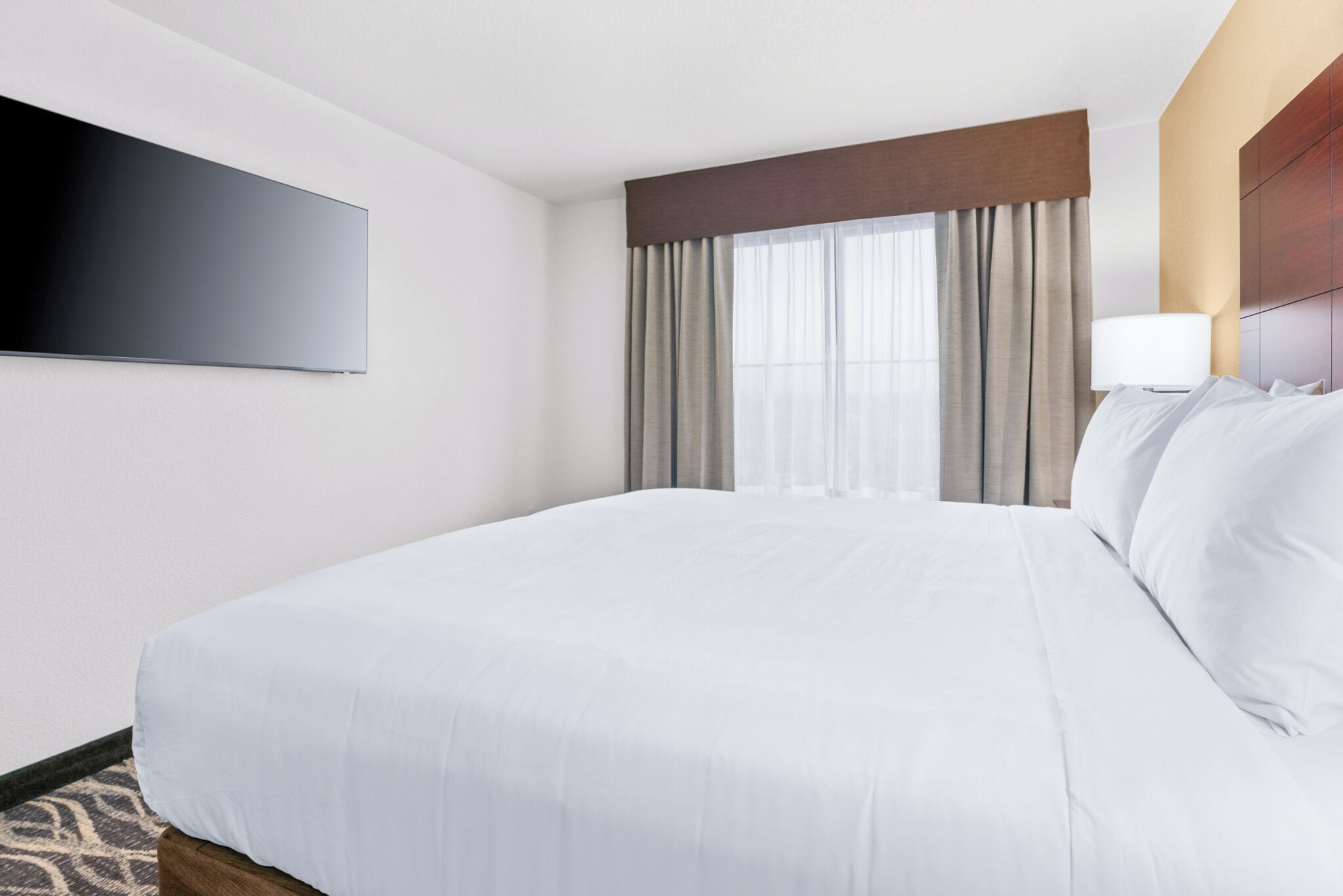 Quarto de hotel com uma cama grande, televisão de tela plana na parede, janela com cortinas e luminária ao lado da cama.