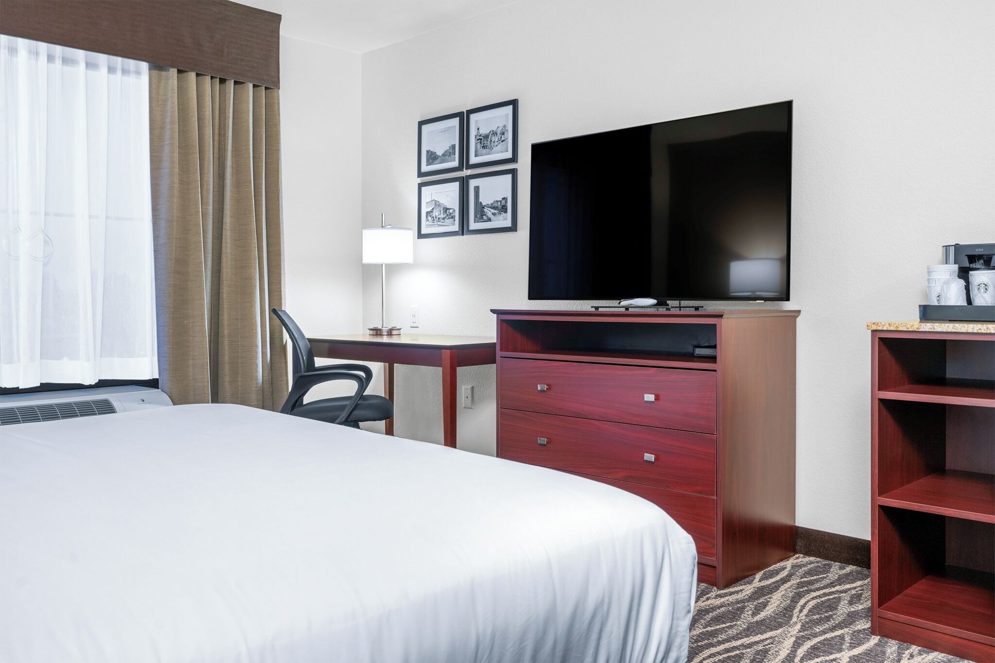 A hotel room with a bed, a desk with a chair, a TV, a dresser, and a coffee machine.