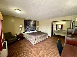 A hotel room with a large bed, a desk with a computer, a chair, a desk lamp, a telephone, and a large mirror.