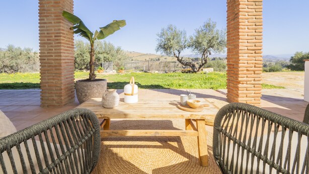 An outdoor patio with a wooden table and chairs, surrounded by brick pillars and a view of olive trees and hills. - Cubo's Casa Rural Pulpito / air conditioning / free wifi / private pool (Coín)