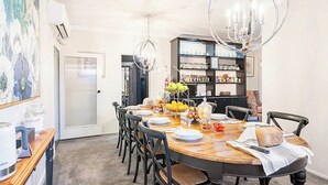 Un comedor con una mesa redonda de madera, sillas negras y una araña. Se observa un área de cocina con estantes y un refrigerador al fondo.