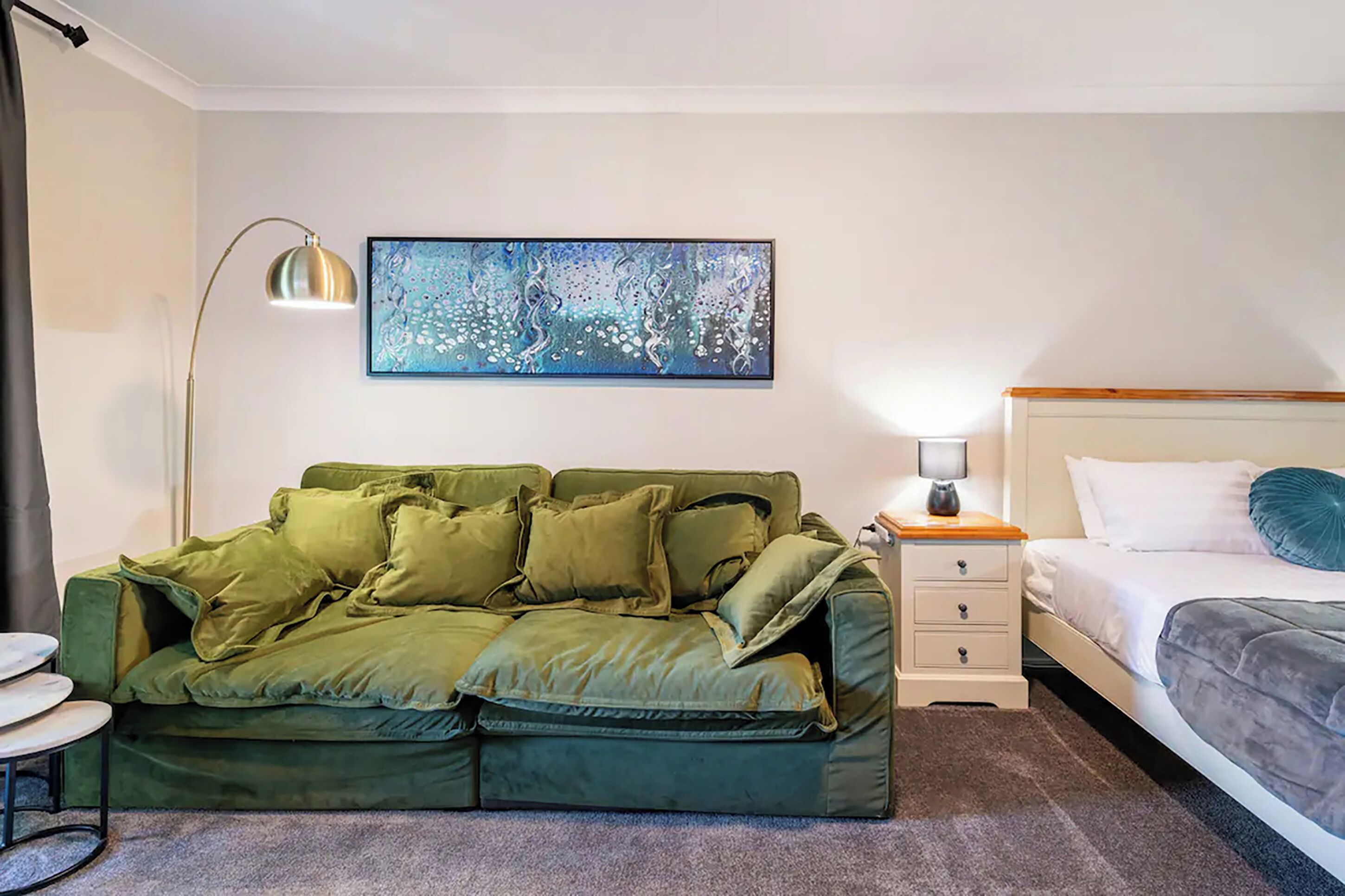 A room with a green sofa, a bed with white bedding, a nightstand with a lamp, and a large abstract painting on the wall.