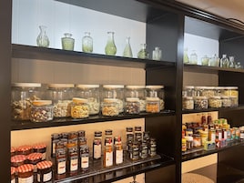Una cocina con estantes llenos de frascos con nueces, cereales y especias, y una encimera con frascos de conservas y condimentos.