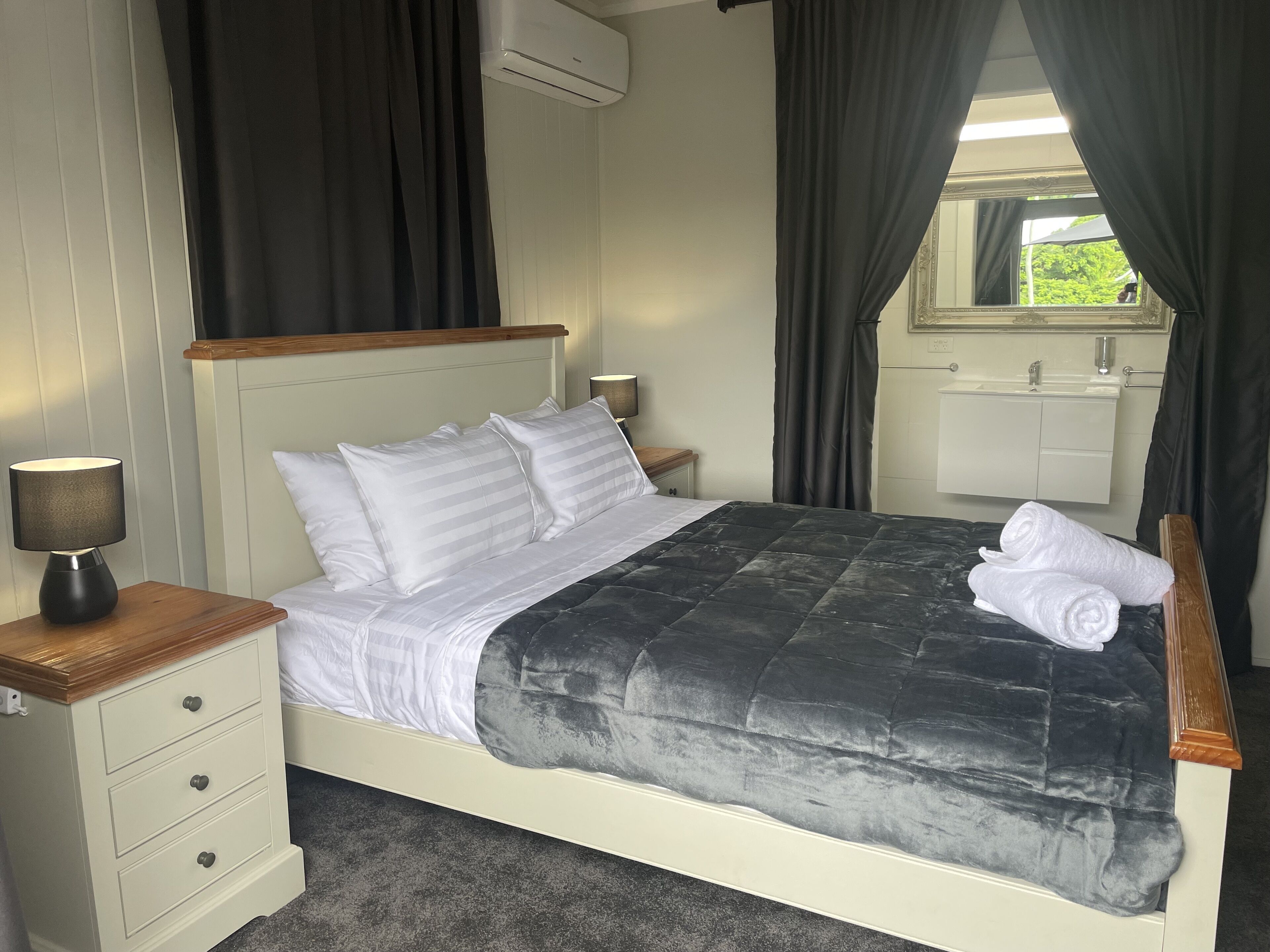 A neatly made bed with white bedding and a dark quilt, a bedside lamp, a wooden nightstand with drawers, and a view of a bathroom through a window with curtains.