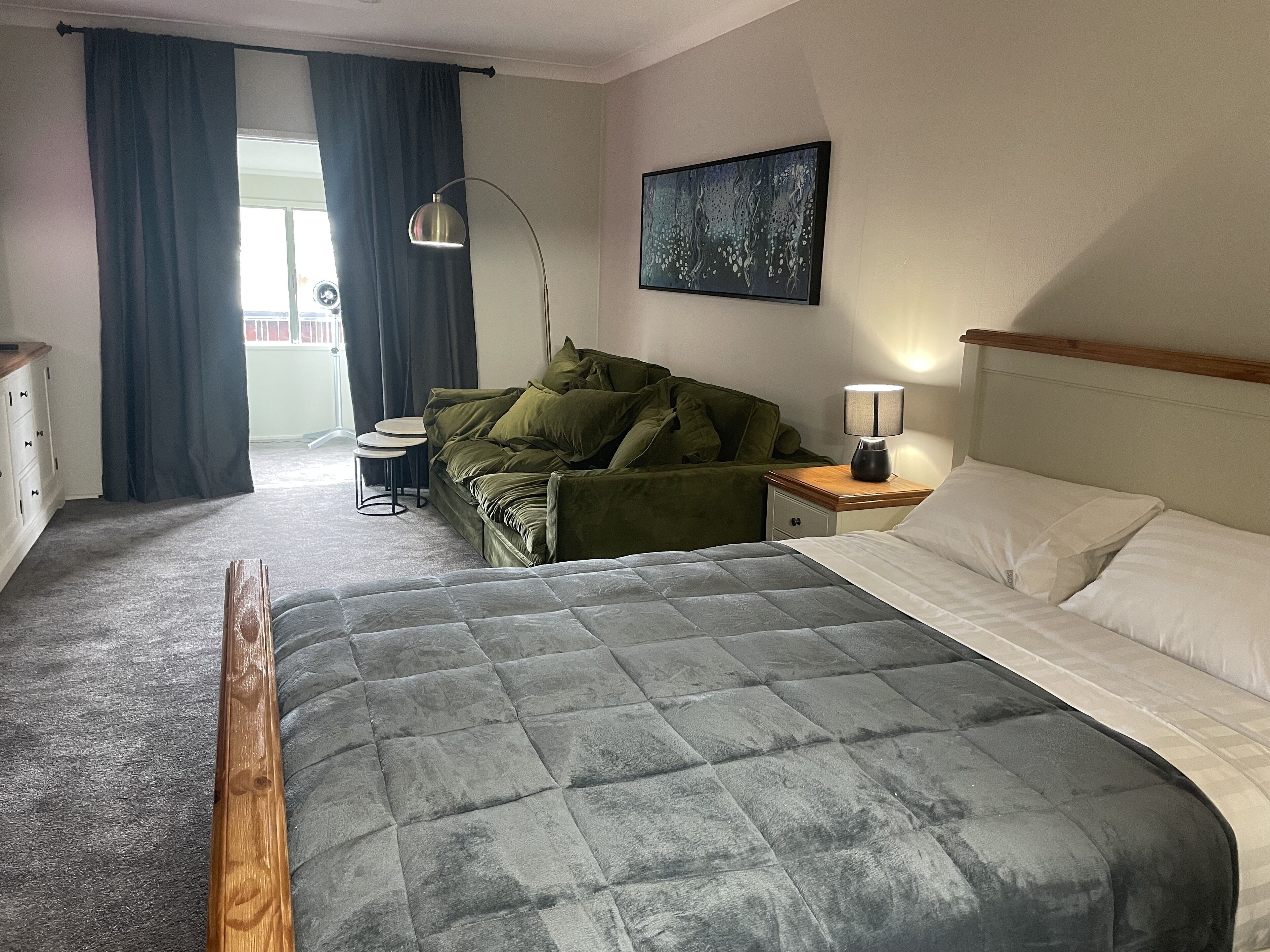 A hotel room with a bed, a green sofa, a small table, a lamp, and a painting on the wall.