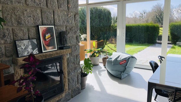 A stone fireplace with framed artwork, a seating area with a grey bean bag, and a view of a garden through large windows. - Jolie Maison Familiale sur la ria à Saint-cado (Belz)