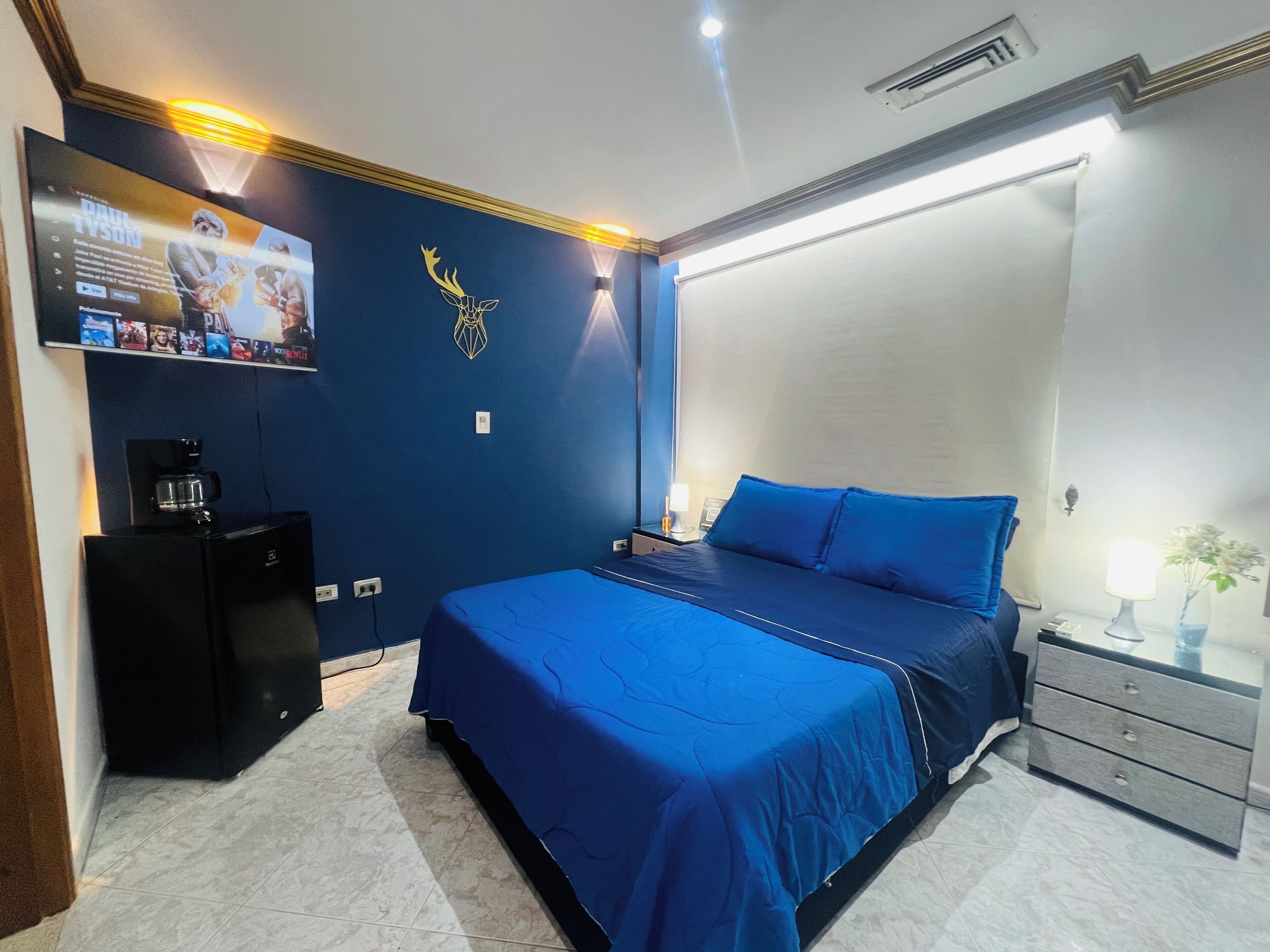 A bedroom with a blue bedspread, a coffee machine, a TV on the wall, and a nightstand with a lamp.