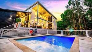 A modern two-story house with a swimming pool, surrounded by trees and a deck with outdoor lighting. - Luxury 3BR in ❤️ of Hochatown! Heated Pool, Playset, Games (Broken Bow)