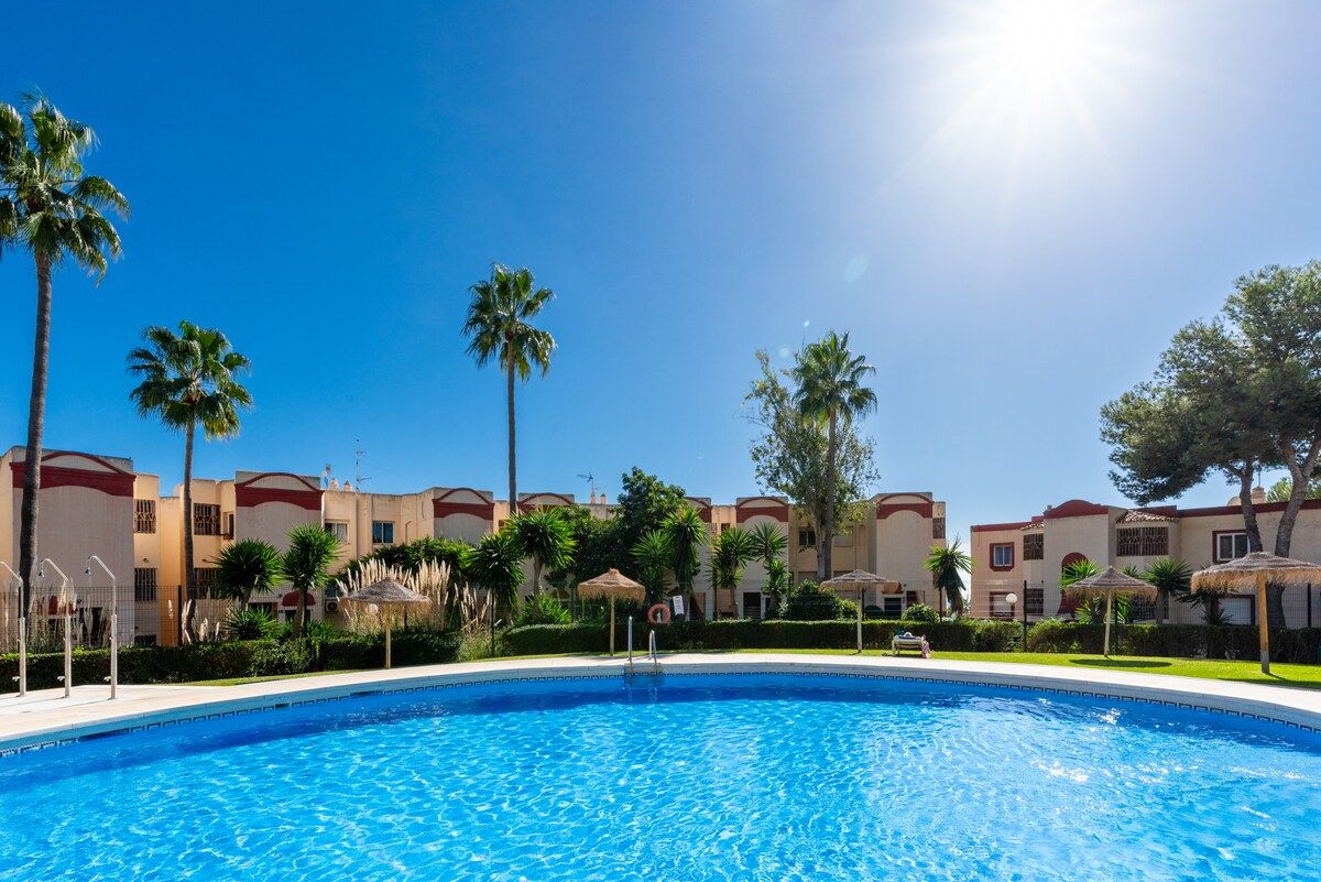 A swimming pool with clear blue water, surrounded by palm trees and a row of buildings with a consistent architectural style.