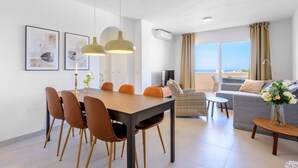 A modern living room with a dining area, a grey sofa, and a large window with a view of the sea.