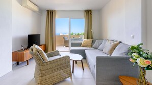 A modern living room with a grey sofa, wicker chair, and a small table. A TV is on a wooden stand, and there's a window with curtains.
