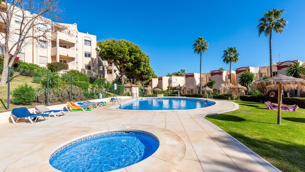 A resort pool area with two circular pools, lounge chairs, palm trees, and a thatched-roof structure.