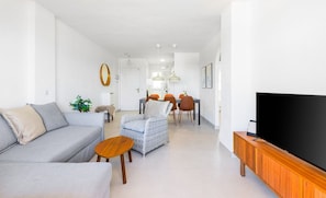 A modern living room with a sofa, a small table, a wicker chair, a dining area with chairs, and a television on a wooden stand. - Zeus | Cosy Apartment with Stunning Sea Views & Pool in Riviera del Sol (Mijas Costa)