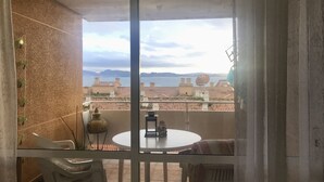 A balcony with a table and chairs, overlooking a coastal cityscape with terracotta rooftops and a body of water.