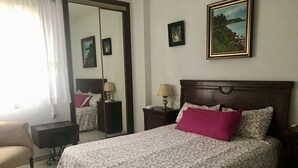 A hotel room with a bed, a chair, a nightstand, a lamp, a mirror, and a painting on the wall.