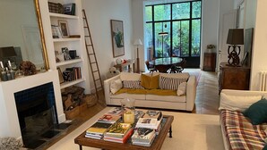 A cozy living room with a fireplace, a white sofa with yellow cushions, a coffee table with books, and a large window with a view of greenery outside.