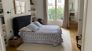 A bedroom with a bed, bedside tables, a bookshelf, a window with curtains, and a door leading to another room.