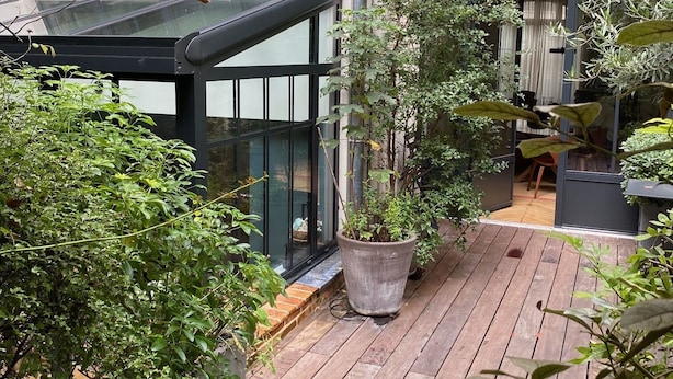 A wooden deck with potted plants, a glass enclosure, and a view of a building with a window.