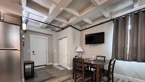 Una cocina funcional con refrigerador, una mesa de comedor con sillas, un televisor montado en la pared y un ventanal con cortinas.